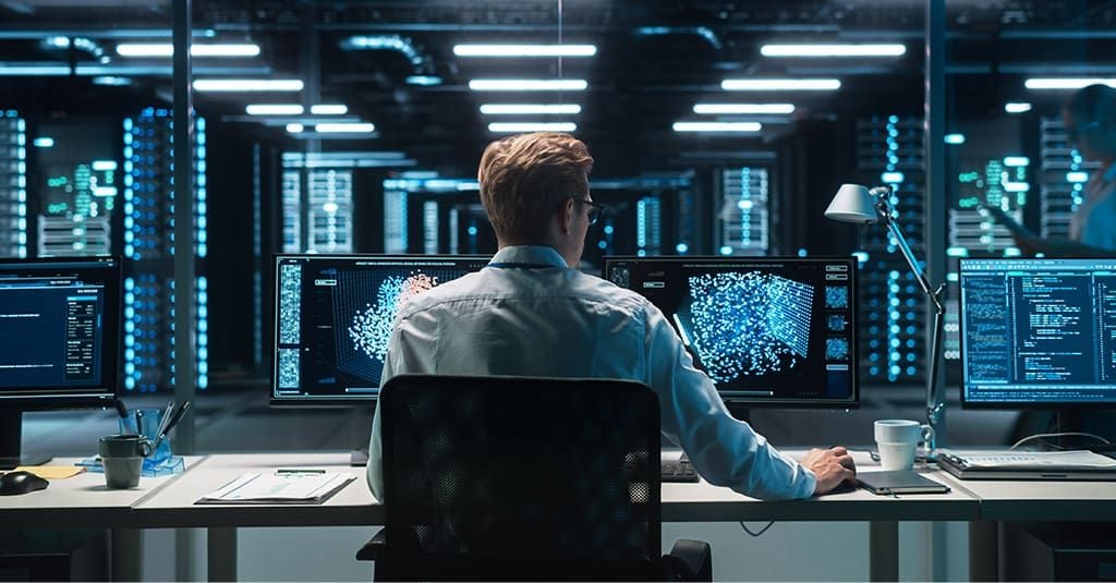 A dark room with many monitors and a man sitting at a desk, looking at them.