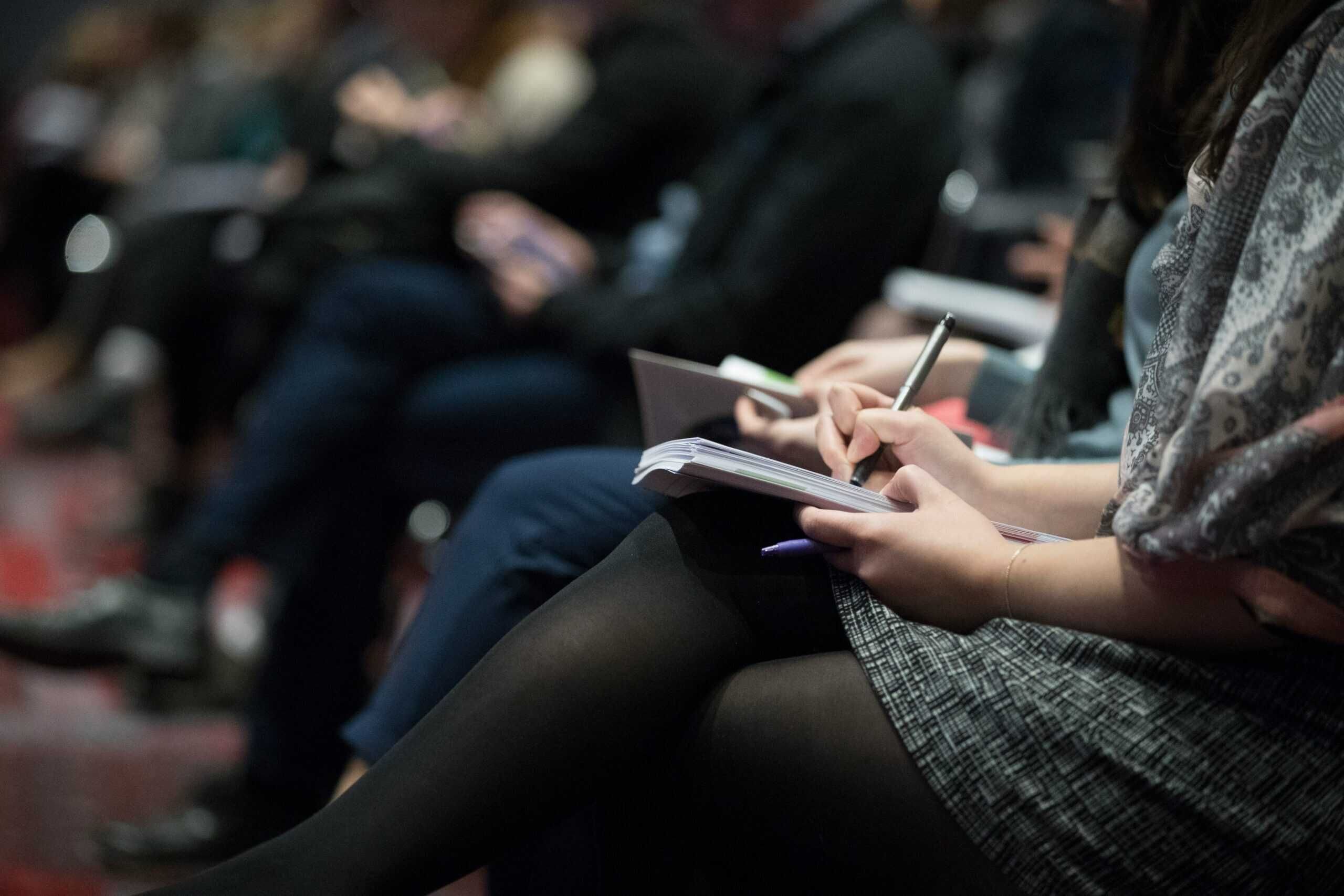 A close-up shot of several people in an auditorium taking notes on their notepads.